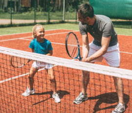 Tennis training. Cheerful father in sports clothing teaching his daughter to play tennis while both standing on tennis court